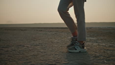 Close-up-shot-of-a-girl-in-gray-shorts-and-sneakers-walking-along-a-deserted-seashore-during-her-vacation-outside-the-city-in-a-summer-evening