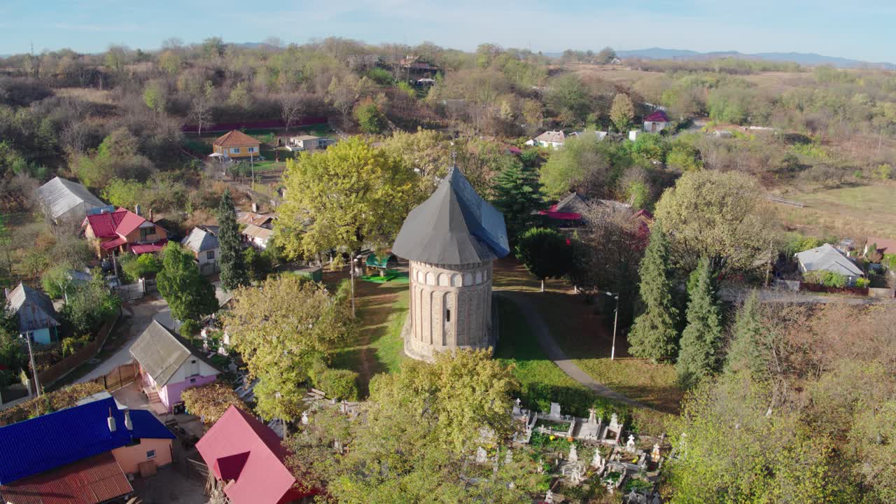A cinematic aerial shot slowly orbits a historic medieval stone church, revealing a cemetery, bright autumn trees, and the surrounding rural village houses in Romania. Stefan the Great, Borzesti