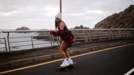 Joven-Despreocupada-Hermosa-Mujer-Hipster-Morena-Con-Abrigo-A-Cuadros-Colgando-De-Las-Manos-Divirtiéndose-Montando-Patineta-Cuesta-Abajo-En-Un-Hermoso-Camino-En-Cámara-Lenta