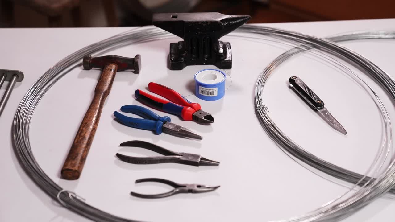 Anvil, hammer, pliers, knife and steel wire prepared on table for traditional Slovak wire craft. Drotarstvo workshop scene showing tools and materials for handmade metal art