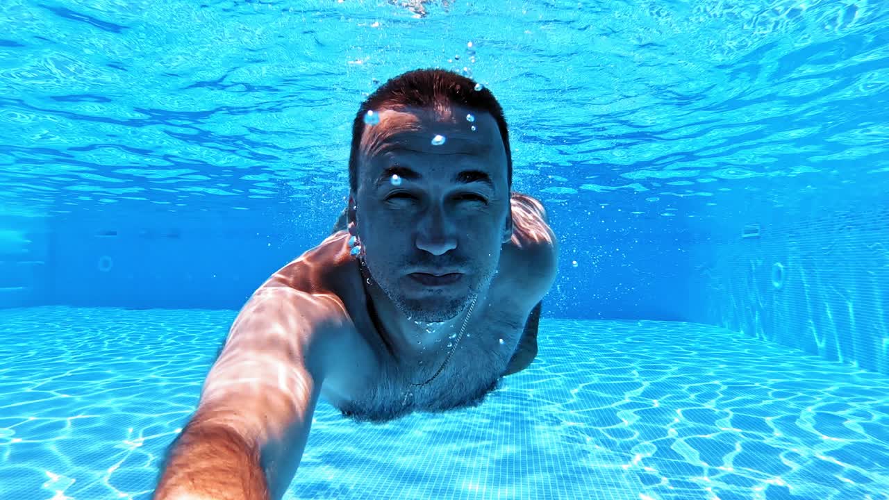 Man swims with underwater camera inside the swimming pool. Transparent water background of a pool and male adult swimming underwater and looking into camera.