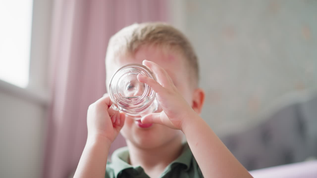 Close up of young kid in green shirt drinking water from transparent glass with soft background and blur view of indoor items suggesting peaceful setting with natural light