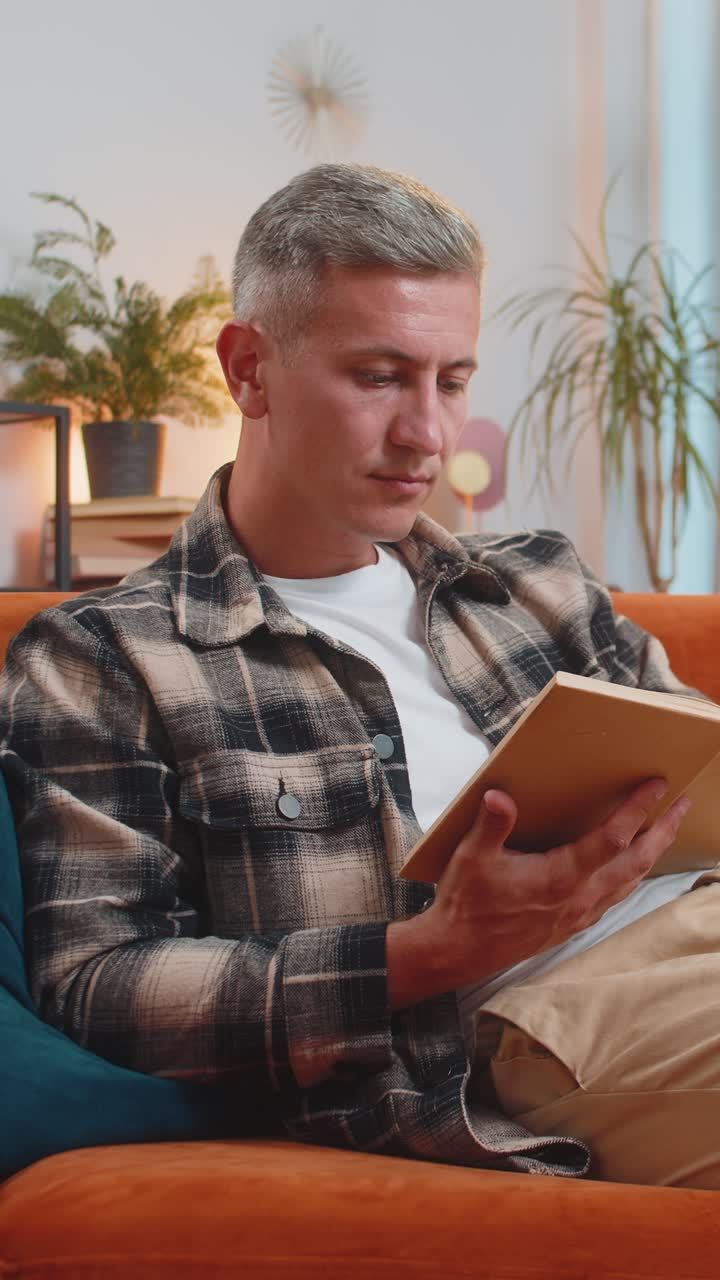 Un homme qui lit un livre intéressant, tourne les pages, sourit, apprécie la littérature, se repose sur le canapé de la maison.