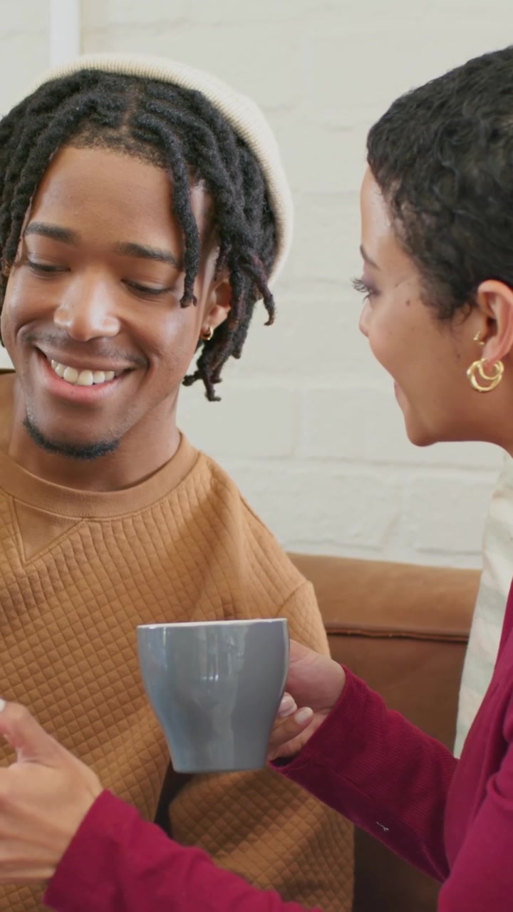 Vertical video: Offering woman leaning toward man holding phone, mug near him on couch talking