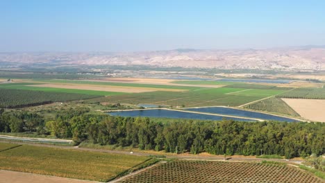 large blue lakes among the green farmlands and the dense forests with the high mountains of northern israel in the background on a sunny day as several cars drive on a road