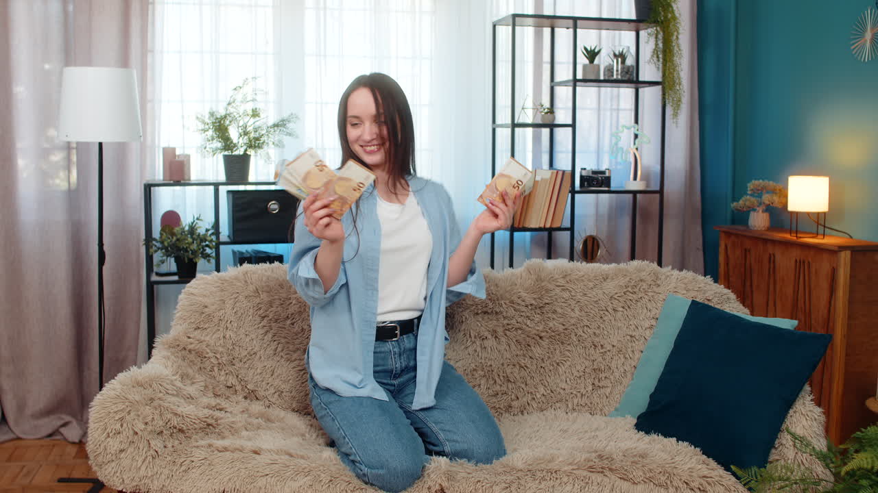 Young woman dancing joyfully on sofa holding cash euro bills celebrating lottery winning success