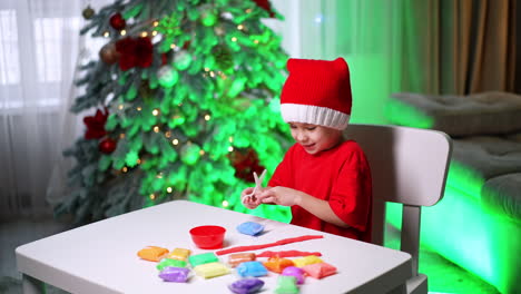 child crafting at christmas time indoors. a child wearing a festive hat sits at a table focused on creating colorful play dough shapes during the holiday season