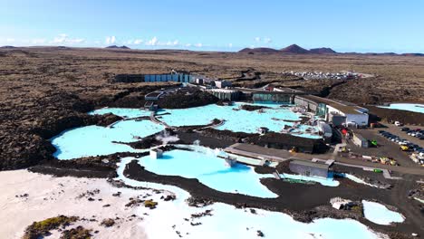 cinematic drone shot of the blue lagoon in iceland, blue sky, desert landscape of lava, sulfur vapor, tourist parking lot