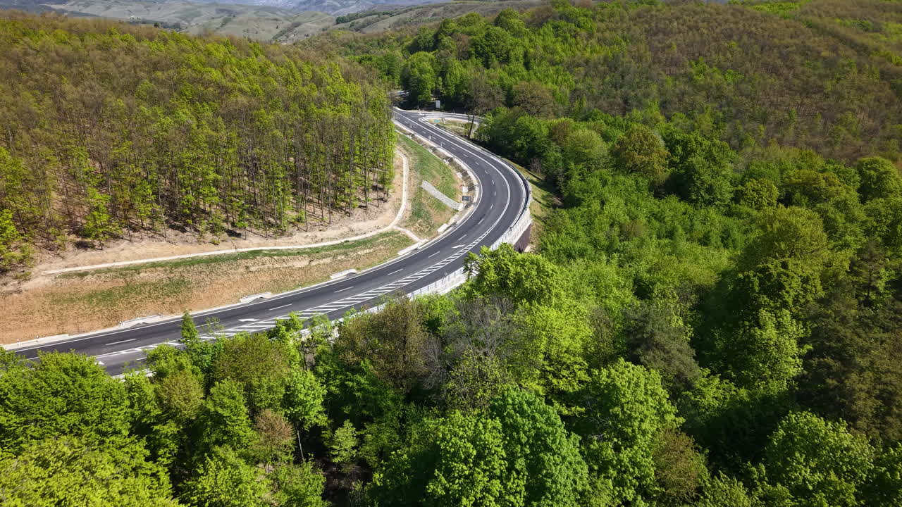 Een luchtfoto toont een kronkelende weg die door een weelderig groen bos snijdt, wat de combinatie van transportinfrastructuur en natuurlijk landschap tijdens de lente benadrukt