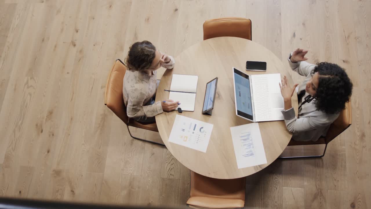 Diverse female colleagues in discussion using laptop and tablet in office lounge, slow motion