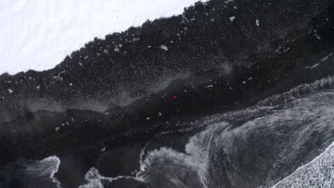 AERIAL:-Overhead-Birdsview-of-Person-walking-on-Black-Beach-with-white-arctic-snow-in-Iceland-in-Winter-Snow,-Ice,-Waves,-Water