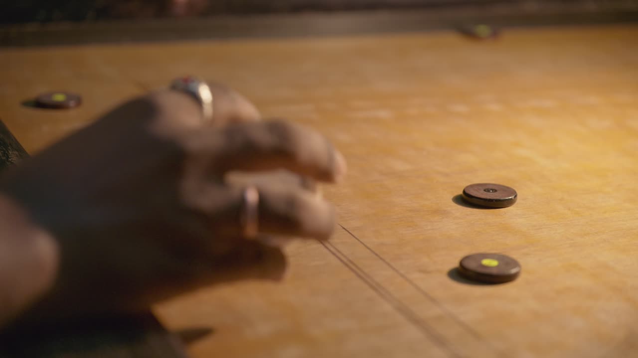 Close-up da mão do jogador atacando o pino no tabuleiro de carrom, moedas deslizando pela superfície de madeira, jogabilidade noturna dinâmica em ambiente interno mostrando velocidade e precisão.