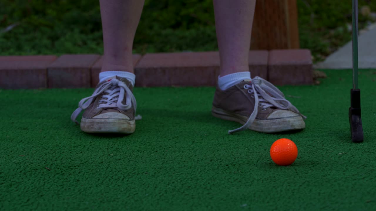 A golfer on a mini golf course placing an orange golf ball on the ground getting ready to shoot the ball