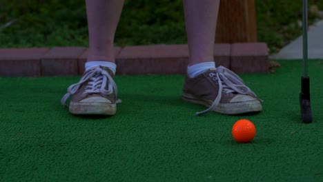 Un-Golfista-En-Un-Campo-De-Minigolf-Colocando-Una-Pelota-De-Golf-Naranja-En-El-Suelo-Preparándose-Para-Disparar-La-Pelota