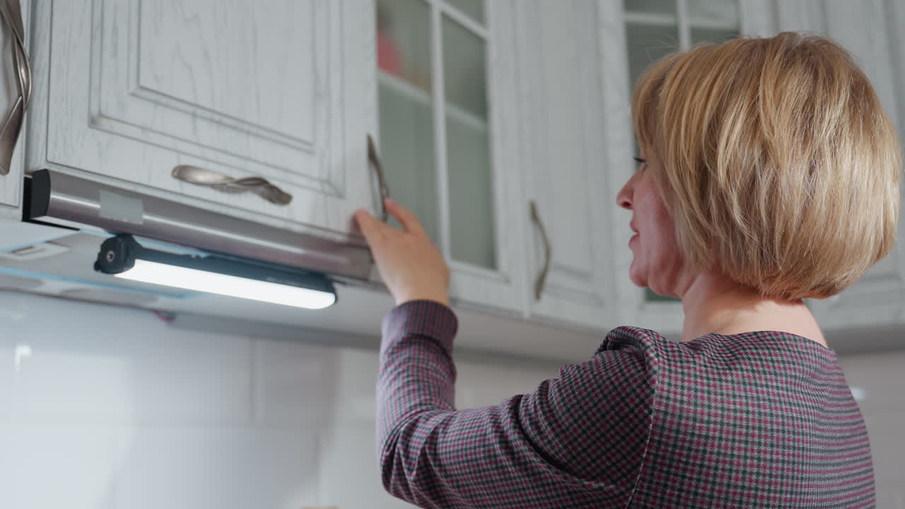 Lady in plaid clothing placing an item inside upper kitchen cabinet while gently closing door, wearing bracelet, modern white kitchen setting with under-cabinet lighting illuminating workspace