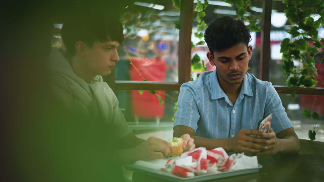European looking man chewing snack while focusing on colleague showing content on phone, casual office break interaction with attentive expression and shared moment over food