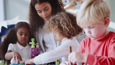 Female-infant-school-teacher-at-table-with-three-children-using-constructing-blocks,-selective-focus