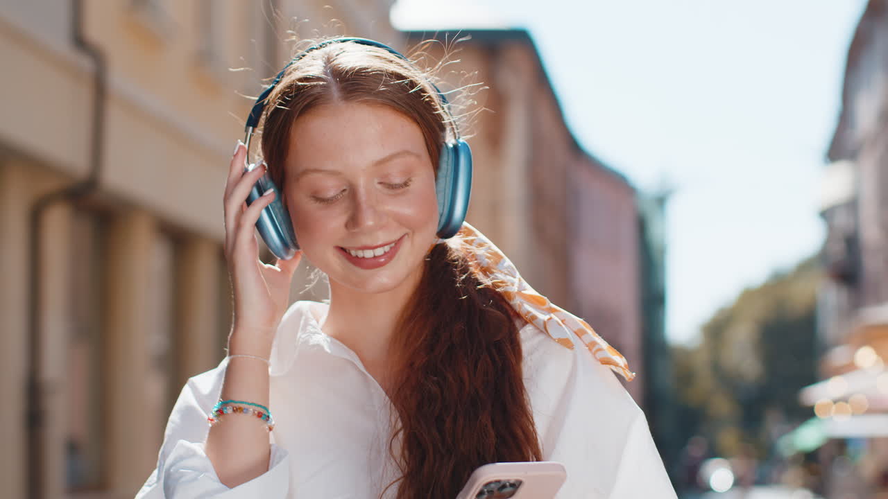 Happy teenager girl in wireless headphones choosing listening music dancing outdoors city street