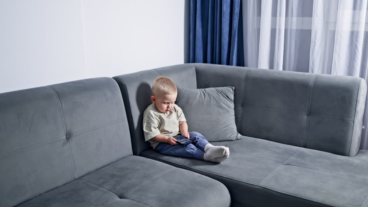 Lille dreng sidder på sofaen derhjemme og leger med telefonen. Barn ser tegnefilm, mens det sidder derhjemme. Tumling ser videoer på telefonen.
