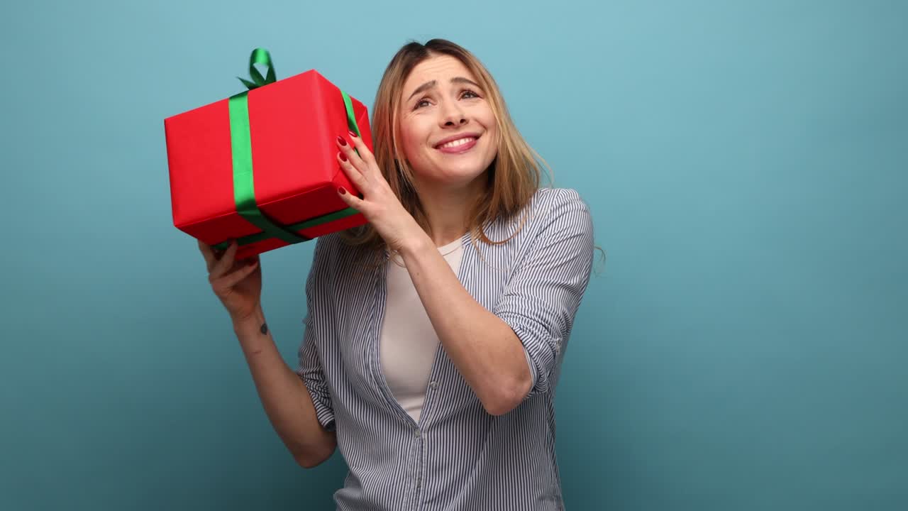 una mujer curiosa de cabello ondulado sacudiendo una caja de regalos envuelta, interesada en lo que hay dentro.