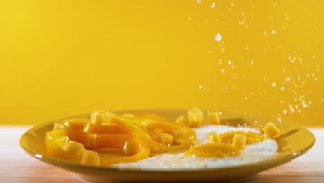 in a slow-motion sequence, salts and vegetable pieces gracefully descend onto a plate with two eggs, set against a yellow backdrop, depicting the art of food preparation and a breakfast scene