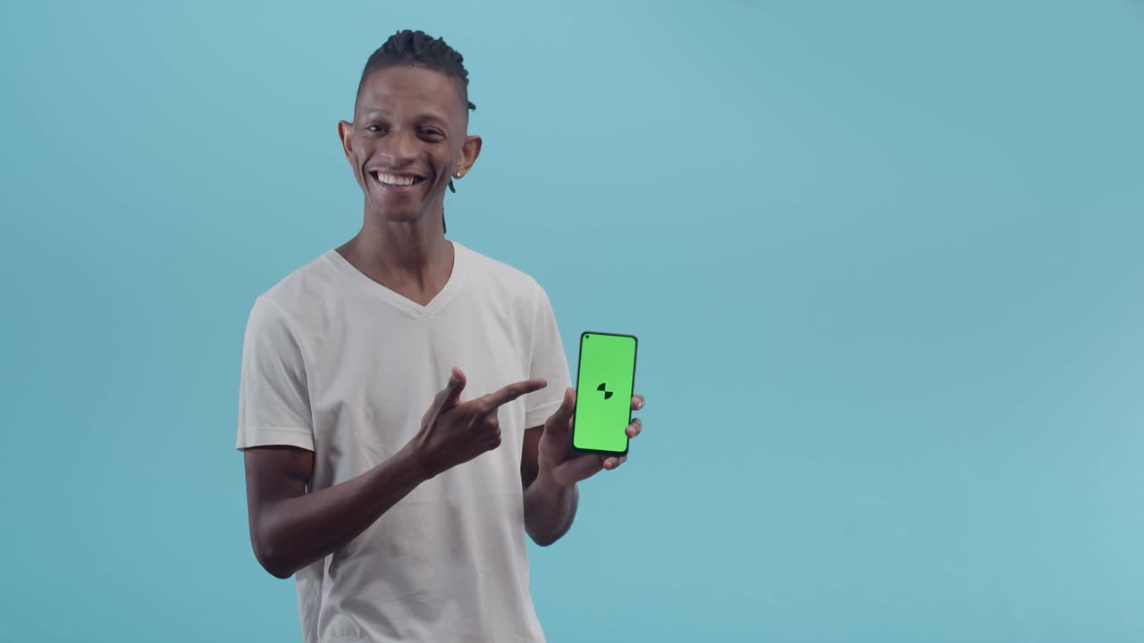 Black man smiles happily showing phone with an app in the studio. Black man with dreadlocks shows a green phone screen standing on a blue background