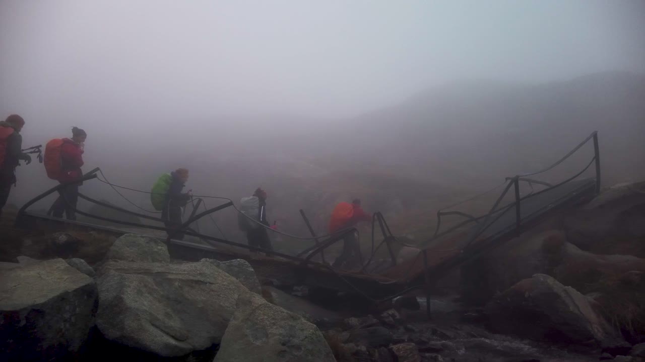 Un gruppo di escursionisti attraversa un ponte rotto su una montagna nebbiosa.