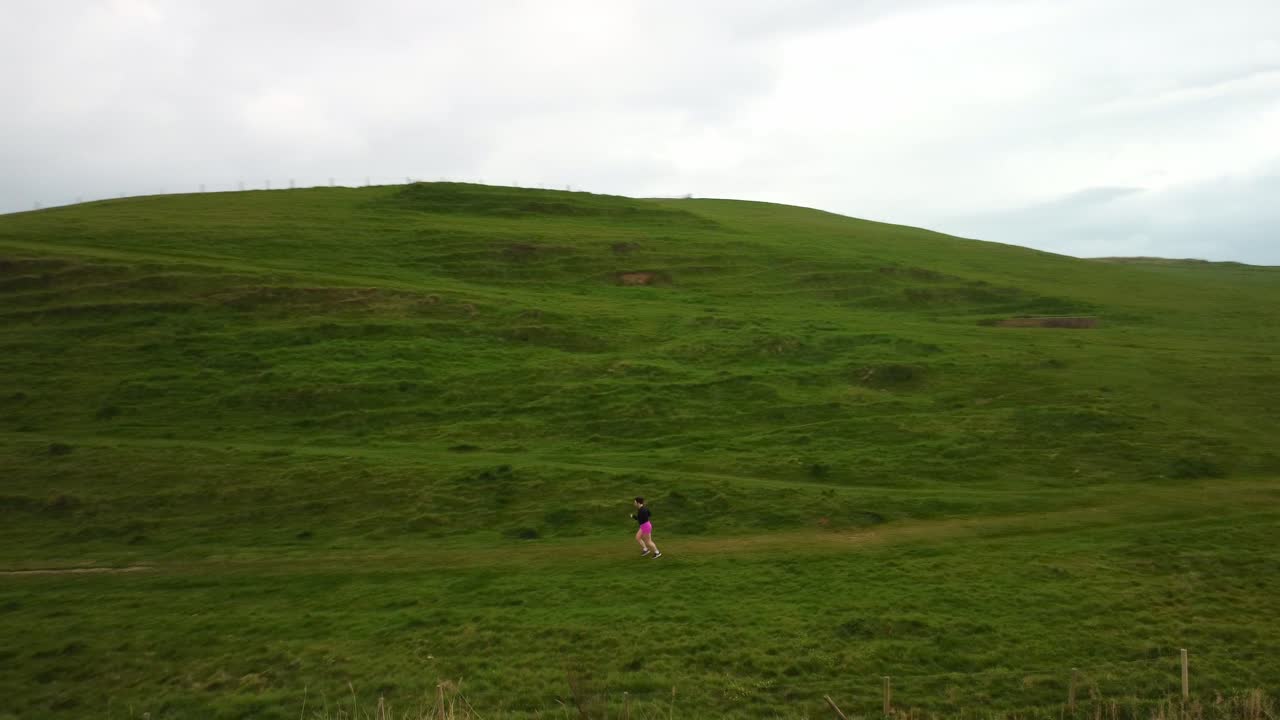 Aerial drone video rotates around a women as she runs along a path in a grassy field. The field is a hill leading up to the top of a cliff that is next to a Jurassic Coast beach. Clouds overhead.