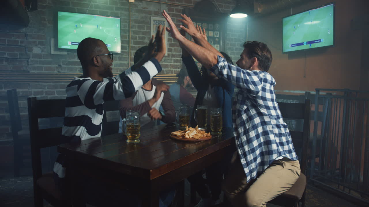 Multiethnic Group Of Friends Sitting At The Bar In The Evening And Giving Five While Watching Sport Game