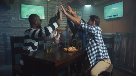 multiethnic group of friends sitting at the bar in the evening and giving five while watching sport game