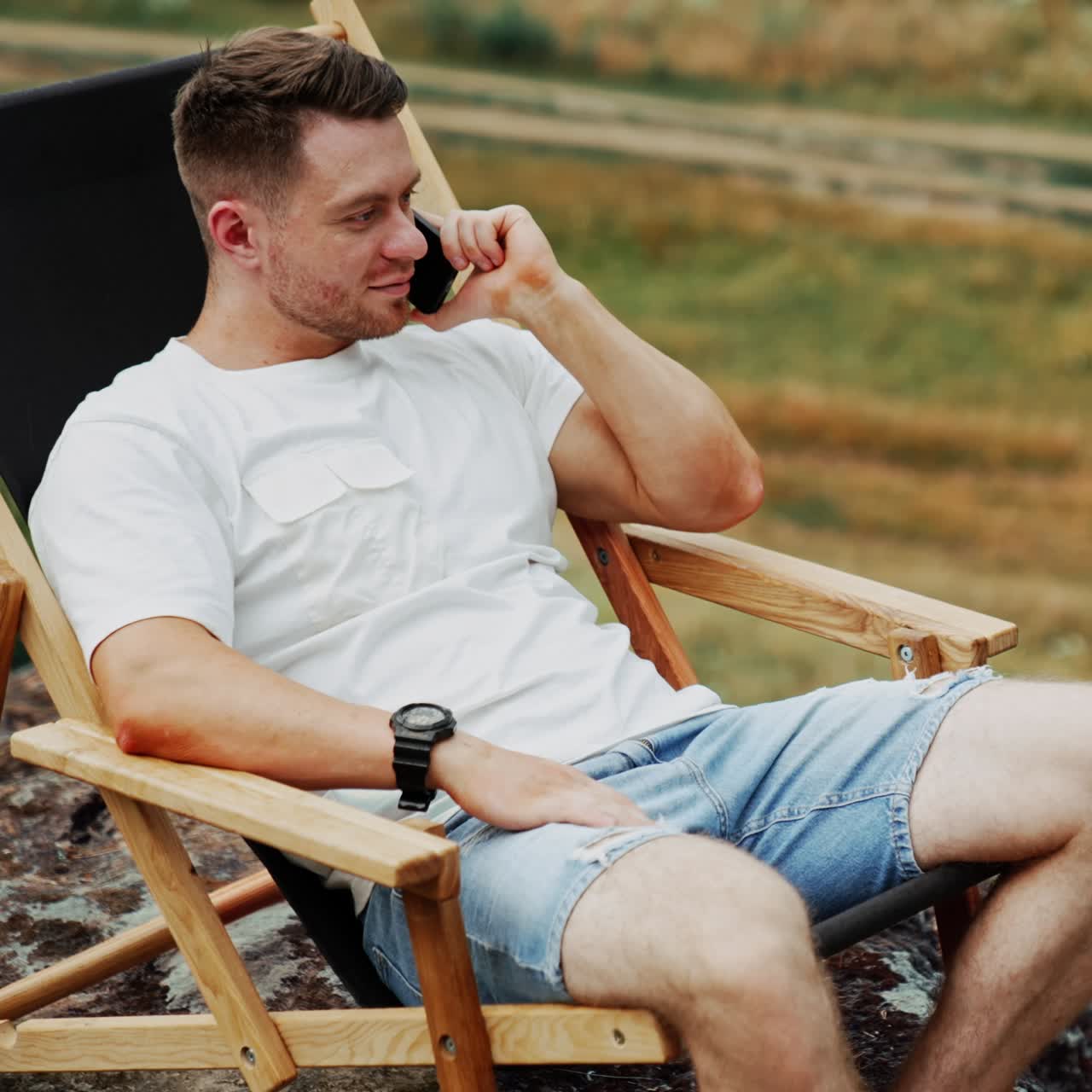 Calm relaxed man wearing white t-shirt and jeans shorts outdoors. Man speaks on the phone sitting in a comfortable folding chair