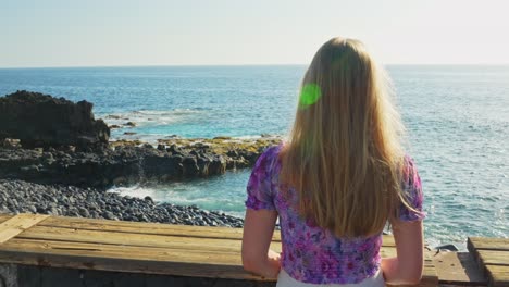 blonde girl standing and looking over calm sea on summer day, rear view