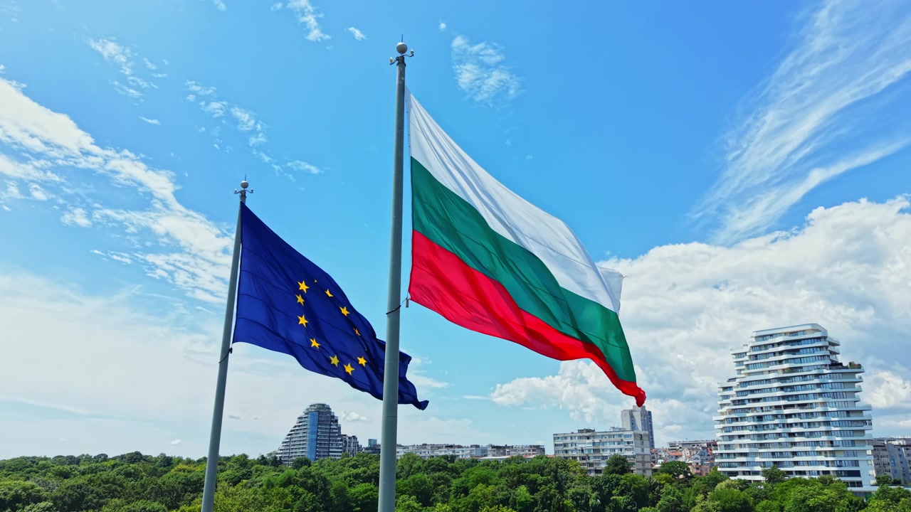 Bandiere della Bulgaria e dell'Unione Europea sventolano nel cielo sopra un paesaggio urbano con edifici moderni durante una giornata limpida