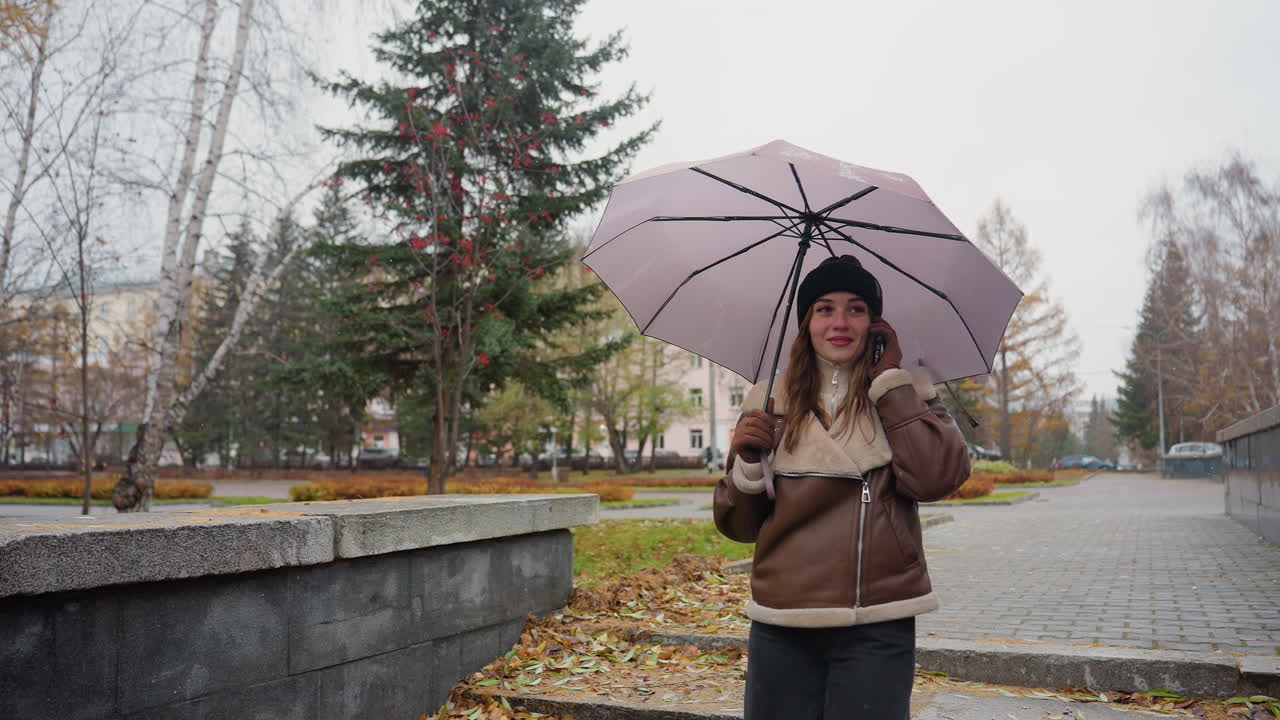 Happy lady smiling on phone call holding umbrella, wearing knit cap, brown shearling jacket, black trousers, walking down stone steps during light snowfall with colorful autumn leaves