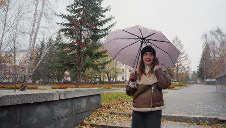 happy lady smiling on phone call holding umbrella, wearing knit cap, brown shearling jacket, black trousers, walking down stone steps during light snowfall with colorful autumn leaves