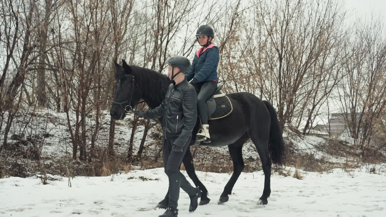 Istruttore che guida un cavaliere con casco a cavallo, un'inquadratura ravvicinata mostra la mano che guida le redini e il passo costante, un mentore paziente che cammina accanto al cavallo scuro, un boschetto innevato e i tetti del villaggio sullo sfondo, equipaggiamento di sicurezza.