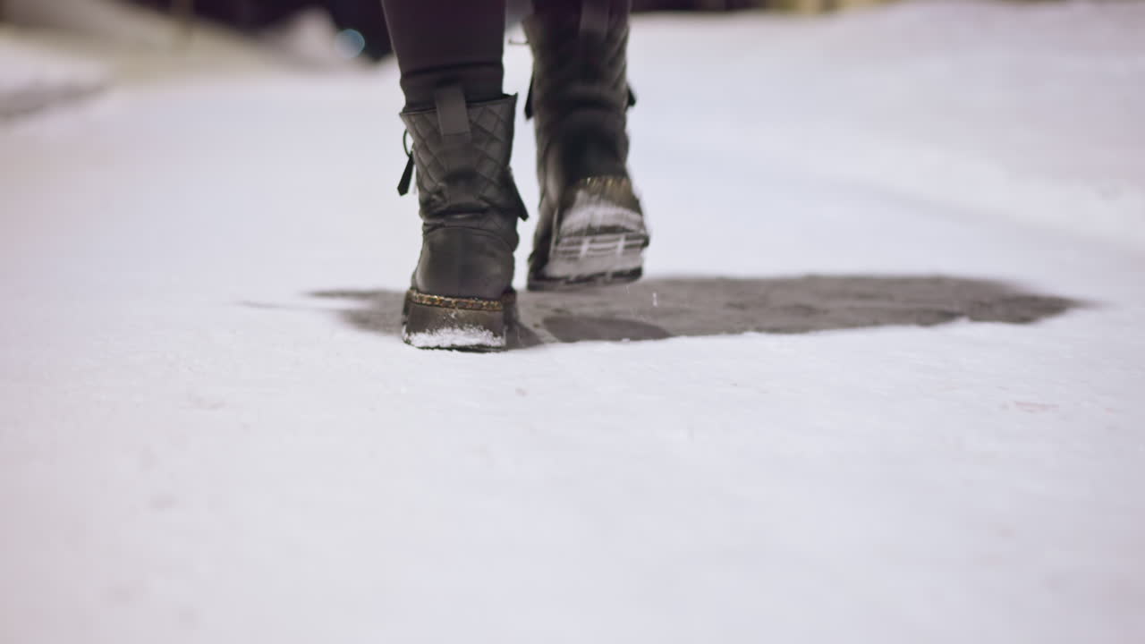 Close up of winter boots walking on snowy path at night leaving footprints, detail of footwear against illuminated ground, person moving through cold season environment under glowing streetlights