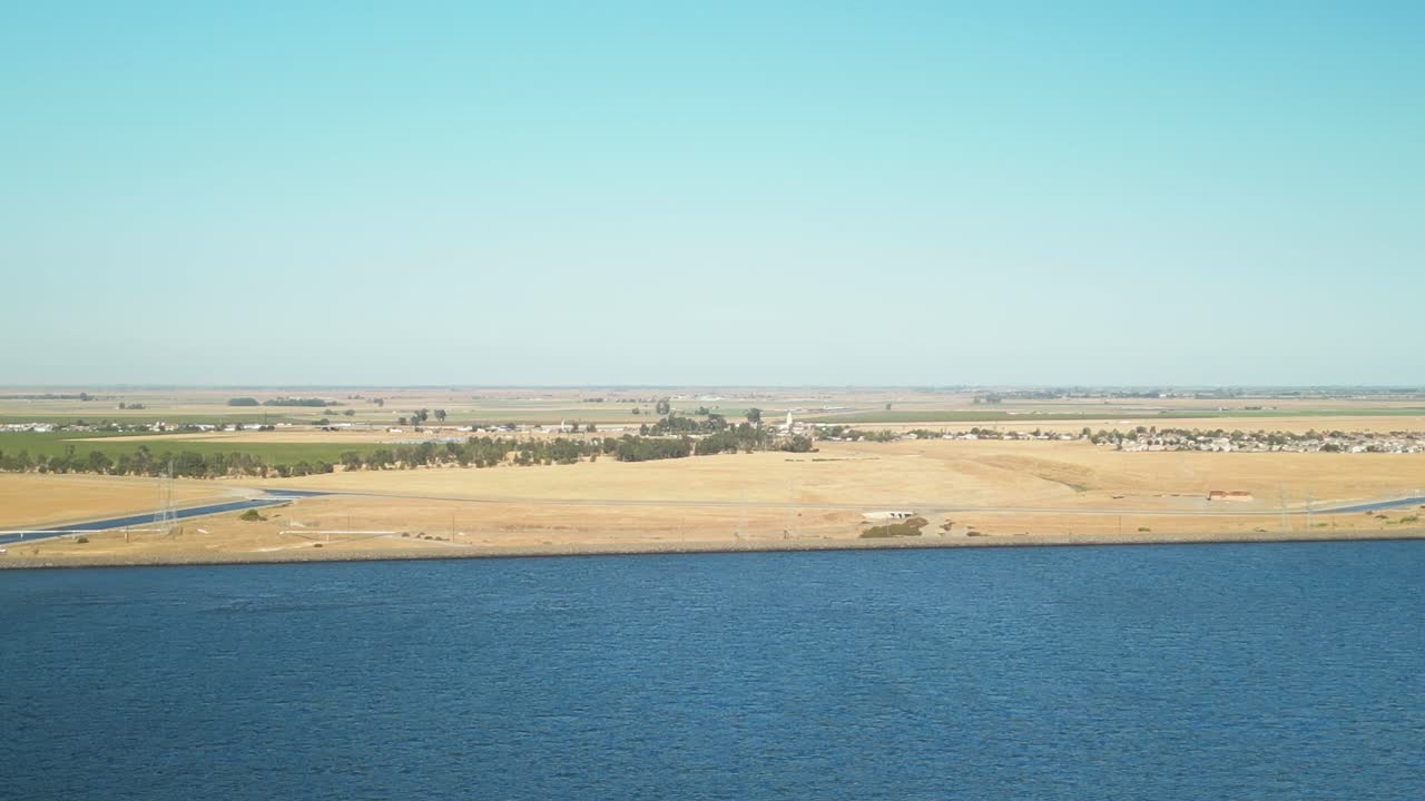 A sweeping drone flight over the shimmering waters of San Luis Reservoir, framed by vast open landscapes and rugged hills under the afternoon sun in Merced County, California, USA.