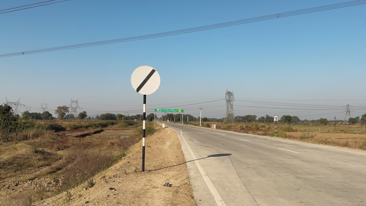 Vídeo de rastreamento gravado ao longo de uma rodovia rural aberta, mostrando uma placa rodoviária indiana indicando o fim da restrição, sinalizando o início dos limites de velocidade nacionais padrão sob um céu azul claro