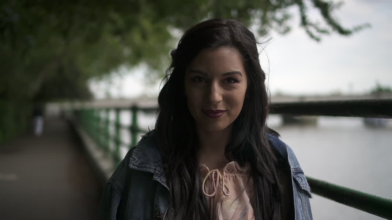 retrato de una atractiva turista latina con cabello negro ondulado y una chaqueta de jean posando en un parque en londres con vista al puente putney detrás de ella