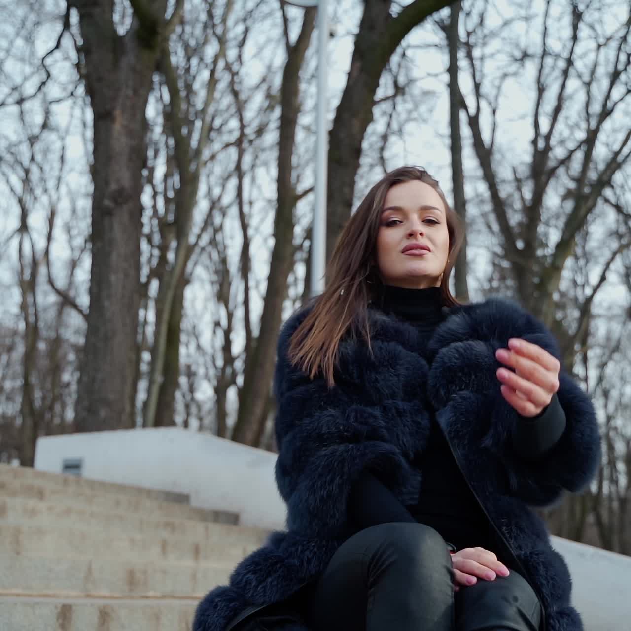 Portrait of attractive model sitting in the park. Beautiful young woman in black fur coat posing on camera outdoors.