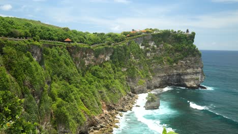 wide shot of the temple on cliff from the other side with waves crashing below, uluwatu, bali