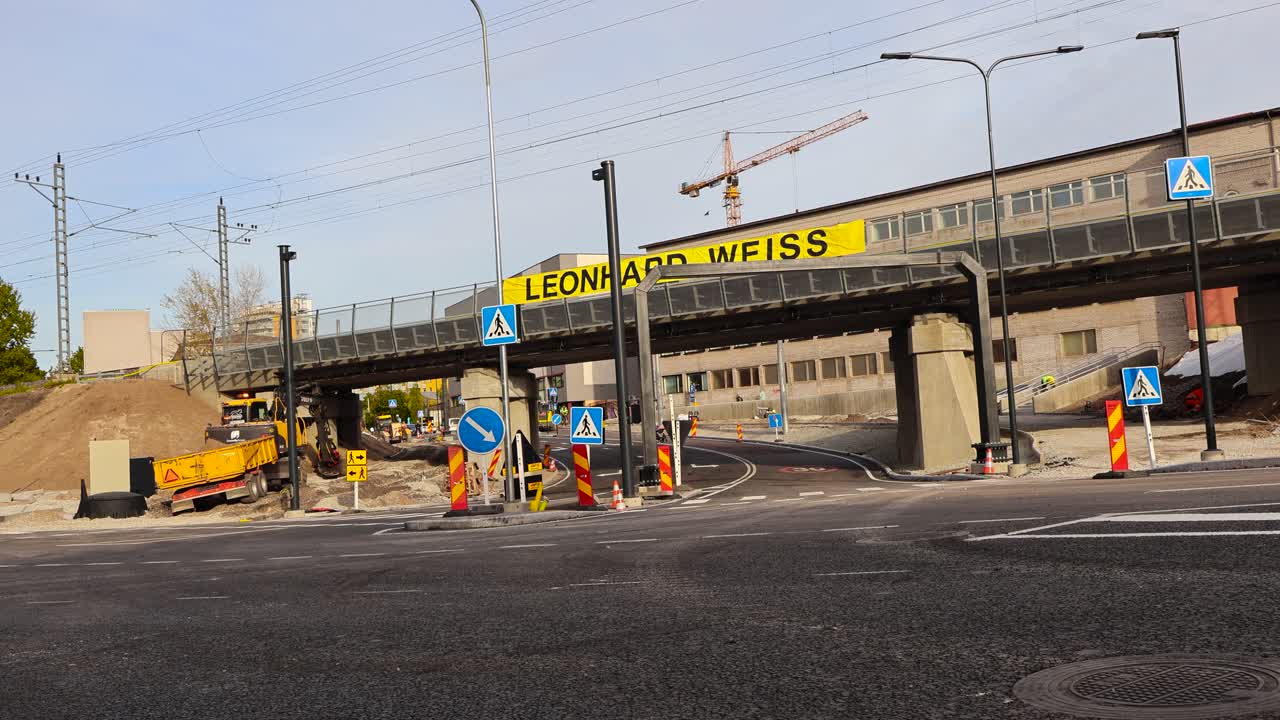 Road construction vehicles, workers and some pedestrians at Tondi bridge under and overpass intersection construction during autumn day time. Temporary traffic signs and new white road markings seen.