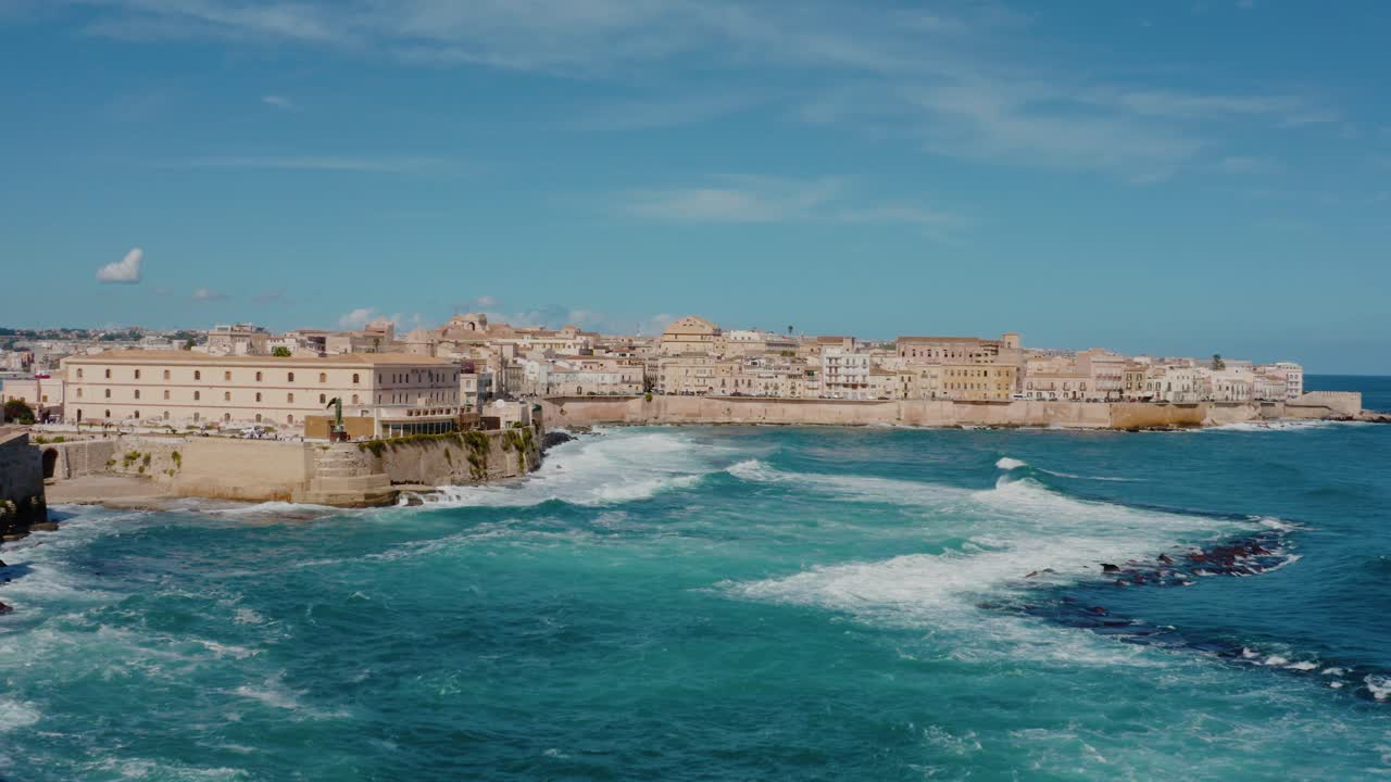 Aerial drone shot of Ortigia, Syracuse. Flying along the coast of Ortigia island with large waves crashing on the shore. Historic Italian town in Sicily.