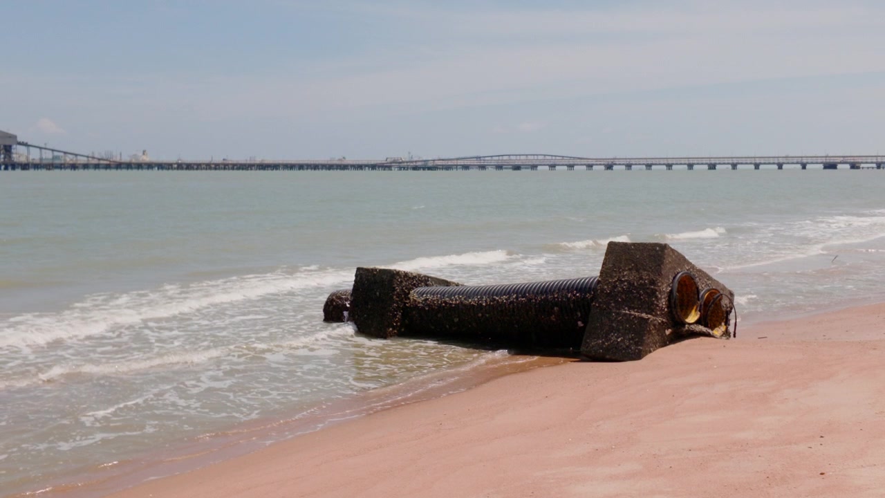 4K slow-motion statische close-up van een verweerde betonnen afvoerpijp op het strand van Pantai Puteri met zachte golven en de verre raffinaderij van Melaka Energy Park, Maleisië