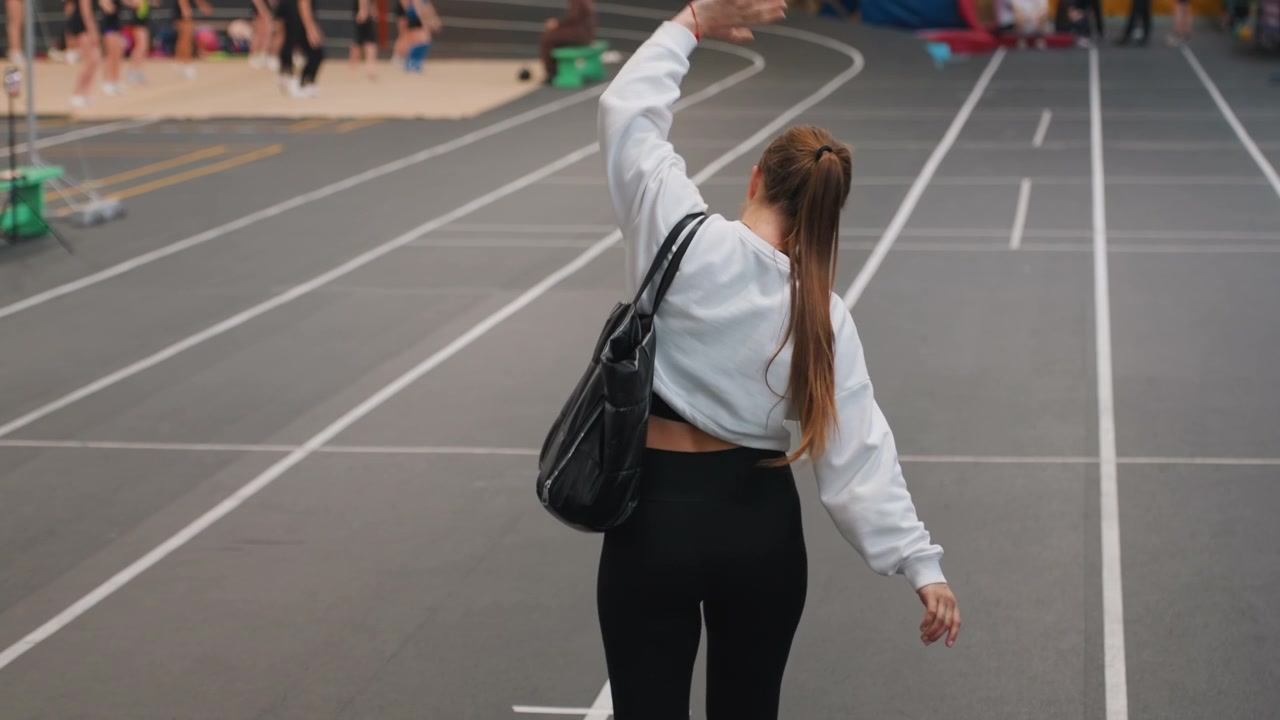 Woman walking away. Braid swings as woman departs scene. Evening lights illuminate woman ending her badminton game. Spectators fade into background as woman with braid and bag exits peacefully