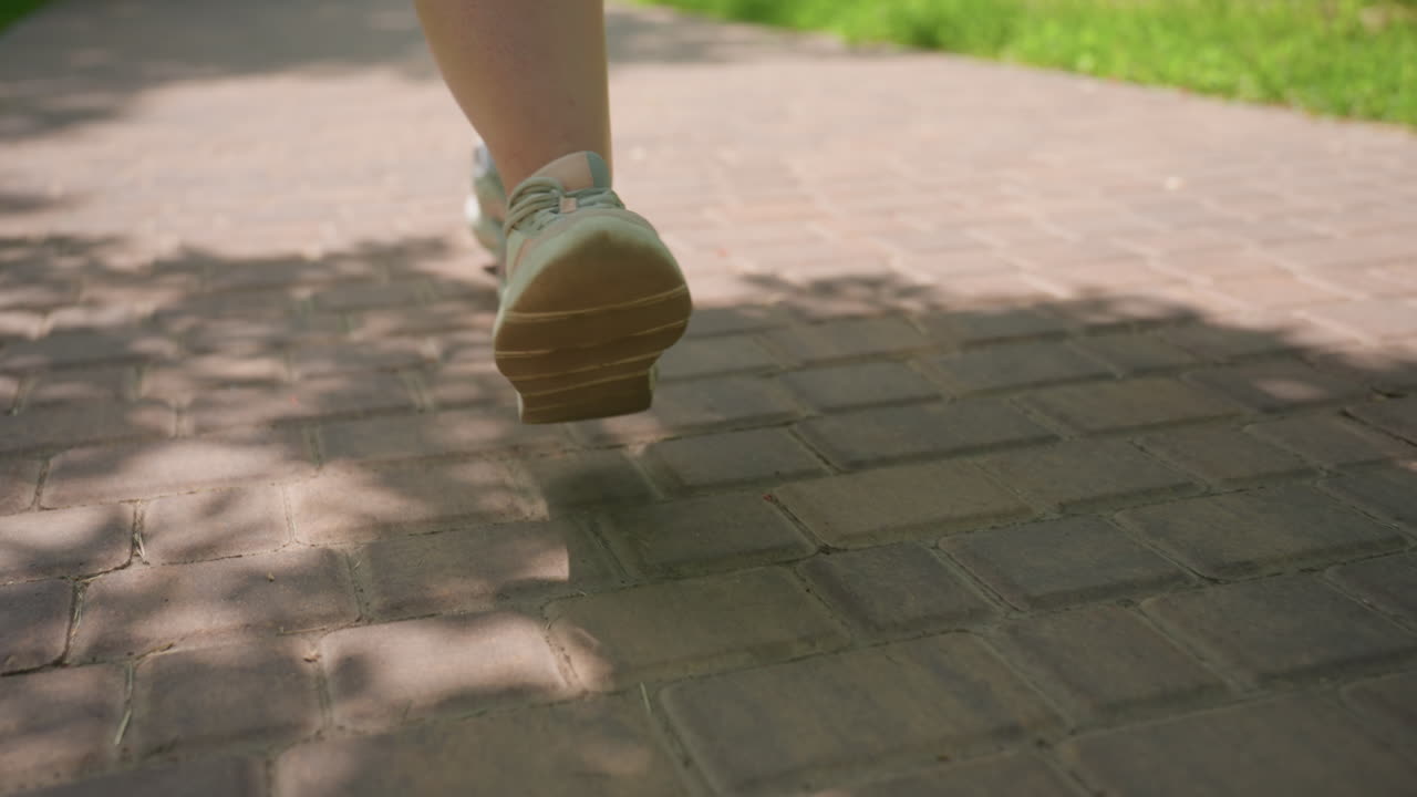 Close up of female sneakers walking slowly on sunny brick pathway surrounded by green grass, peaceful summer day atmosphere, soft motion capturing leisure walk