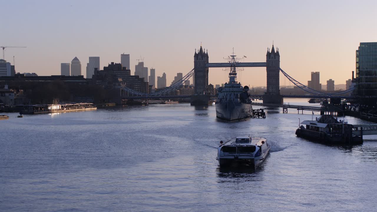 Wide shot of a cruise boat leaving the pontoon on the River Thames with Tower Bridge and the London skyline in the background, bathed in warm evening light.