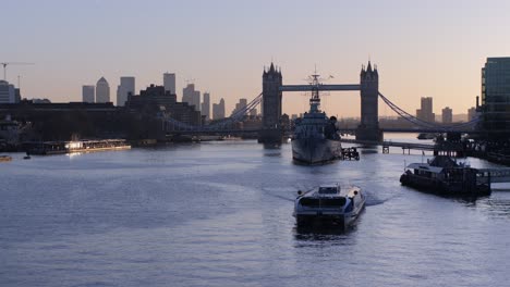 wide shot of a cruise boat leaving the pontoon on the river thames with tower bridge and the london skyline in the background, bathed in warm evening light.