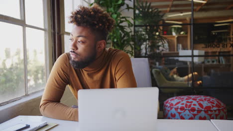 millennial black male creative sitting at a desk in an office, looking out of the window, close up